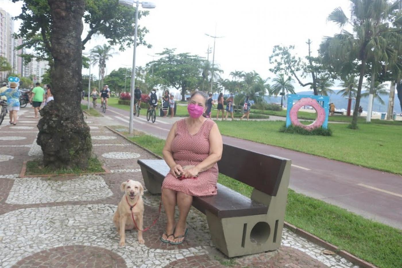 mulher está sentada no banco da praia acompanhada de um cachorro. Ao lado direito, a ciclovia e o jardim. #paratodosverem