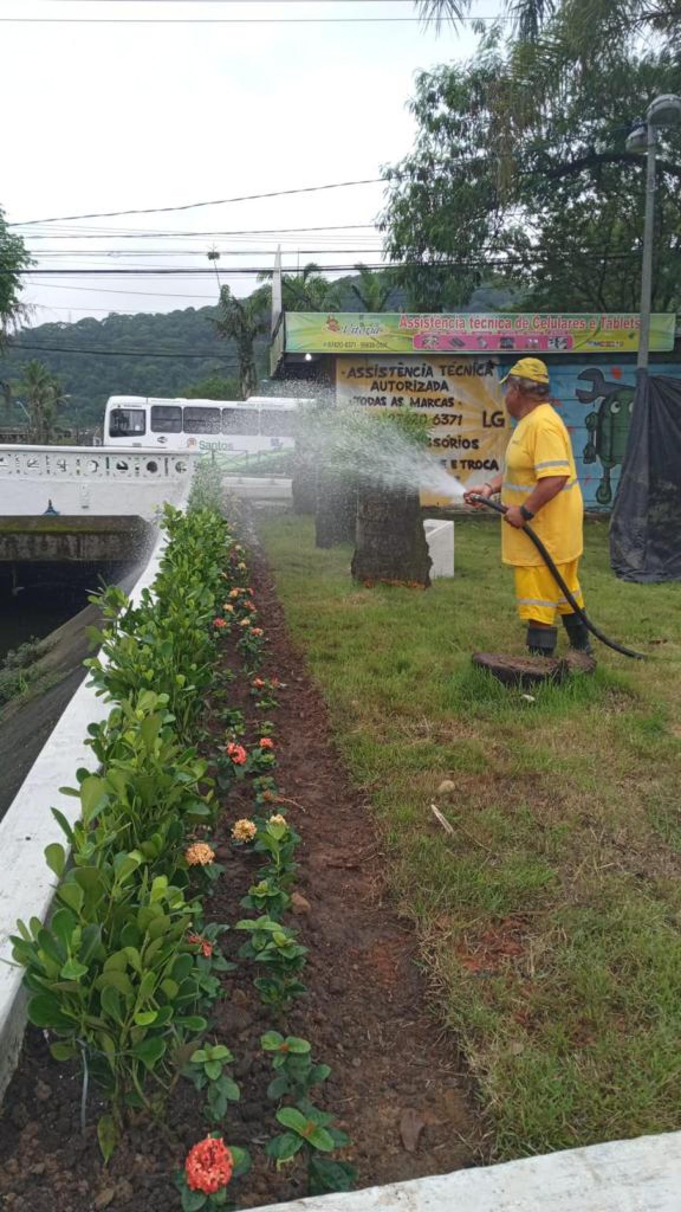 homem uniformizado esguicha água de mangueira em canteiro com flores à margem de um canal. Ele está sobre a grama. #paratodosverem