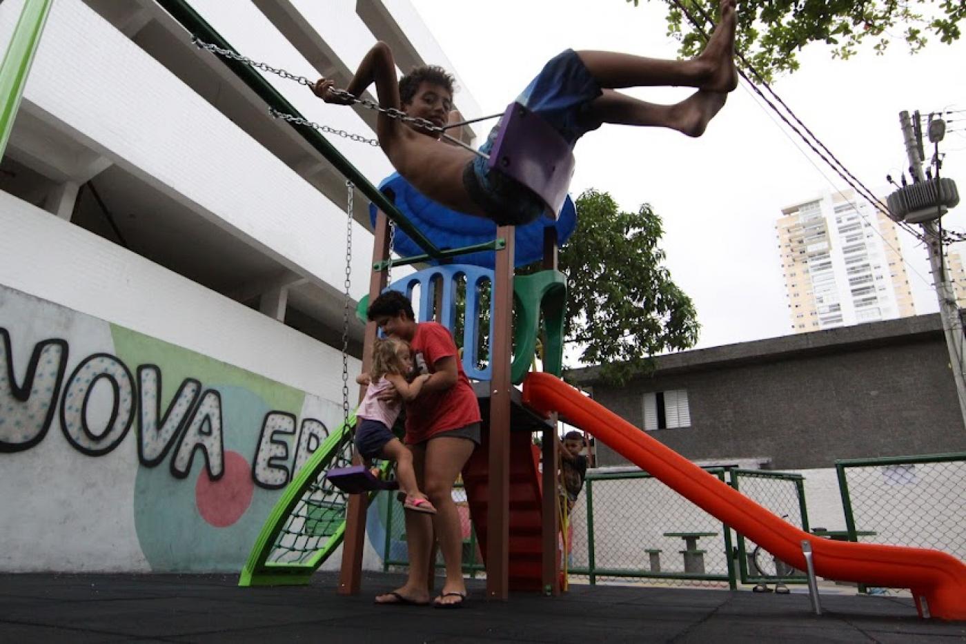 criança está brincando no balancê. Ao fundo, um escorregador com uma mulher segurando uma criança. #paratodosverem