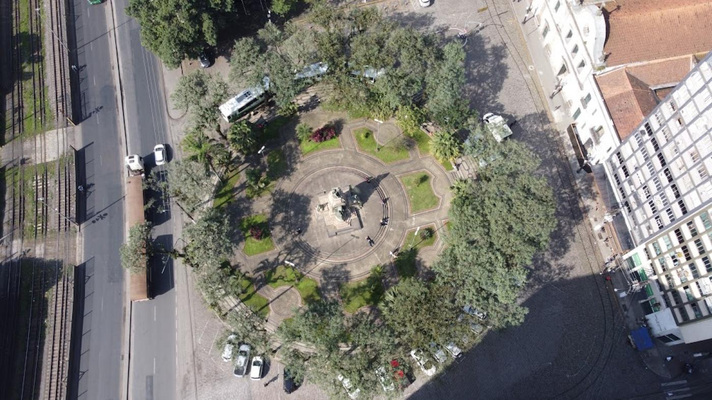 vista aérea da praça com avenida do lado esquerdo, onde passa um caminhão, e rua ao lado direito. A praça é cercada por árvores. #paratodosverem