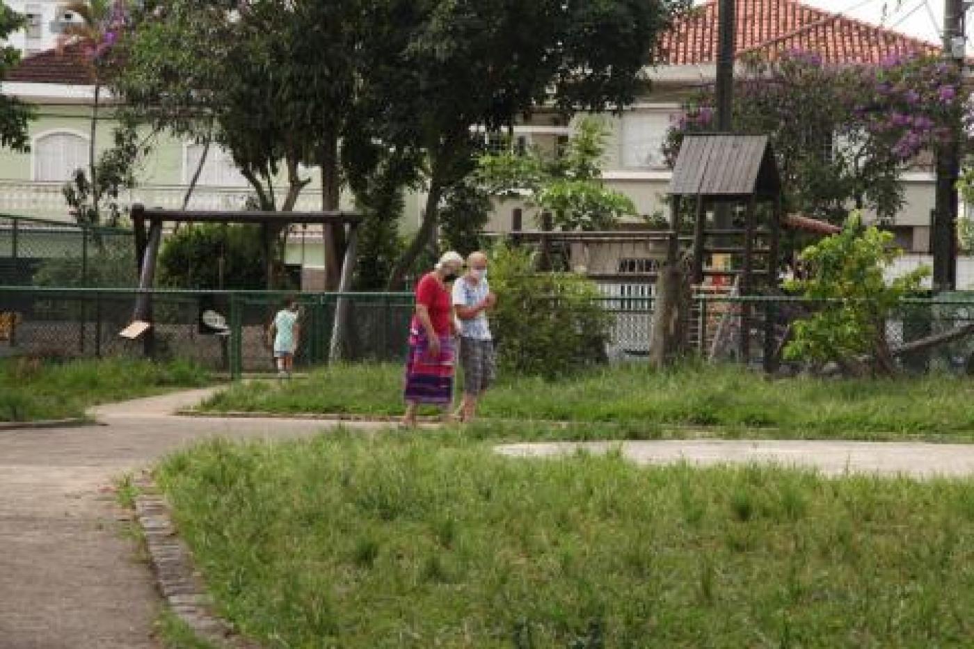 praça com área gramada. Dois idosos caminham. Um playground ao fundo. #paratodosverem