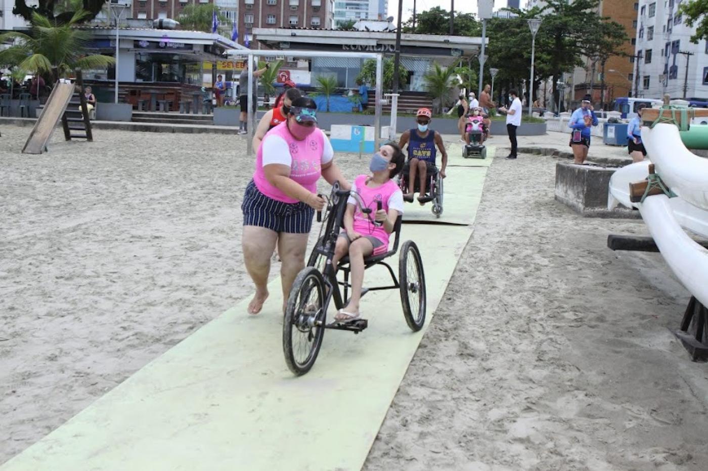 Jovem anda de triciclo, guiada por mulher em pé, em tapete sobre a faixa de areia. #pracegover