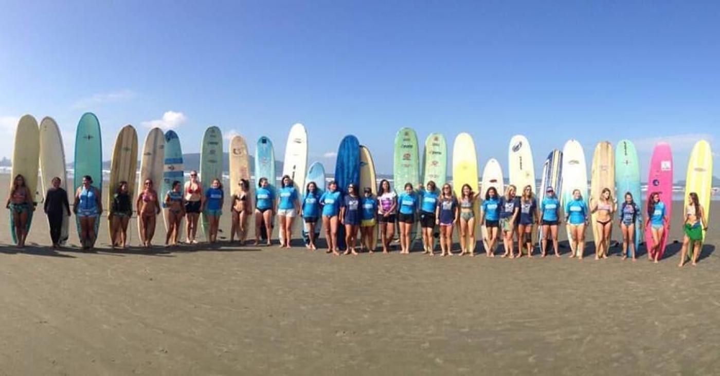 Várias mulheres em pé na areia à frente de seus respectivos long-boards. #Pracegover