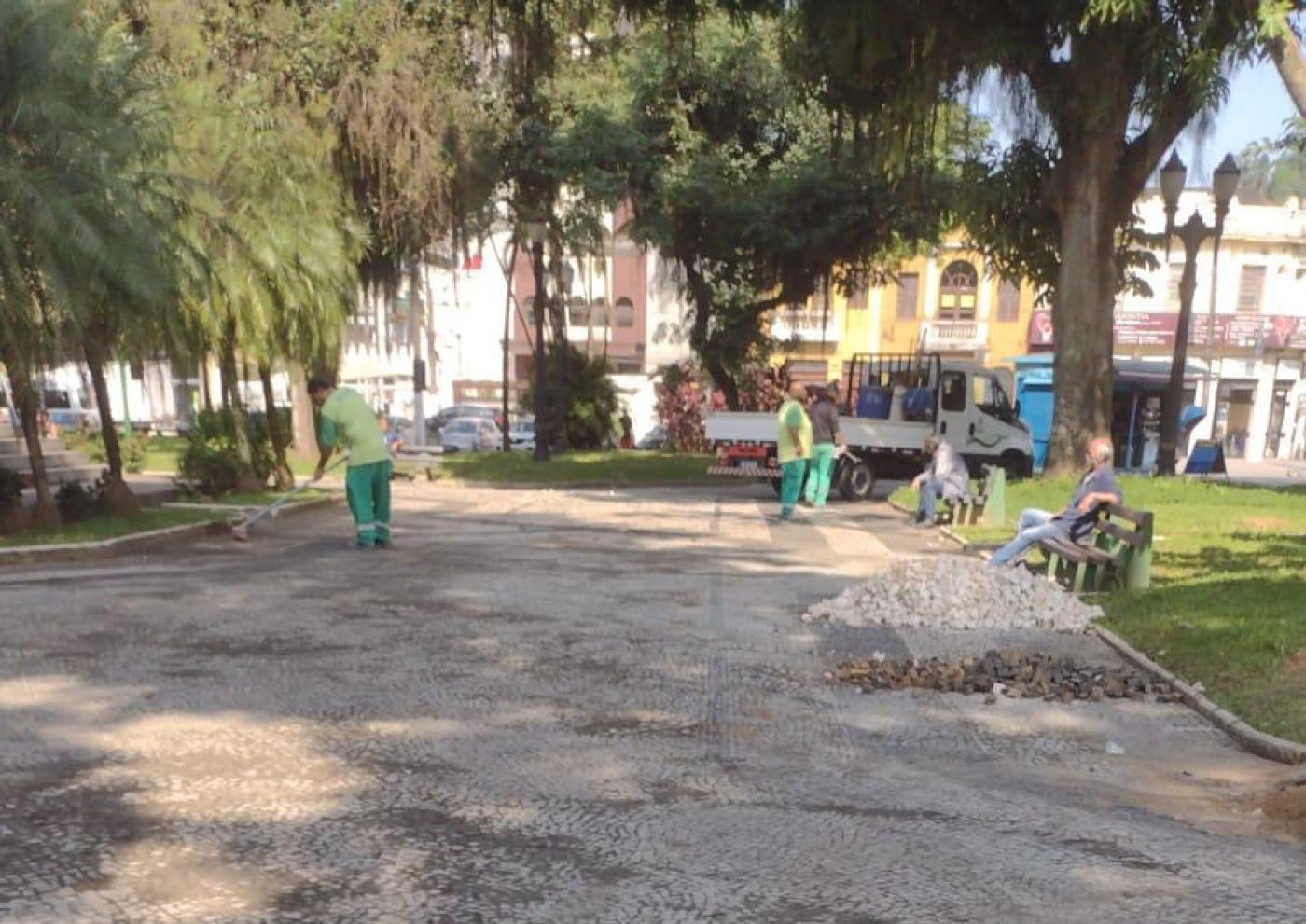 homens estão trabalhando em praça. Eles estão uniformizados. Há pedras de mosaico português em um canto. #paratodosverem