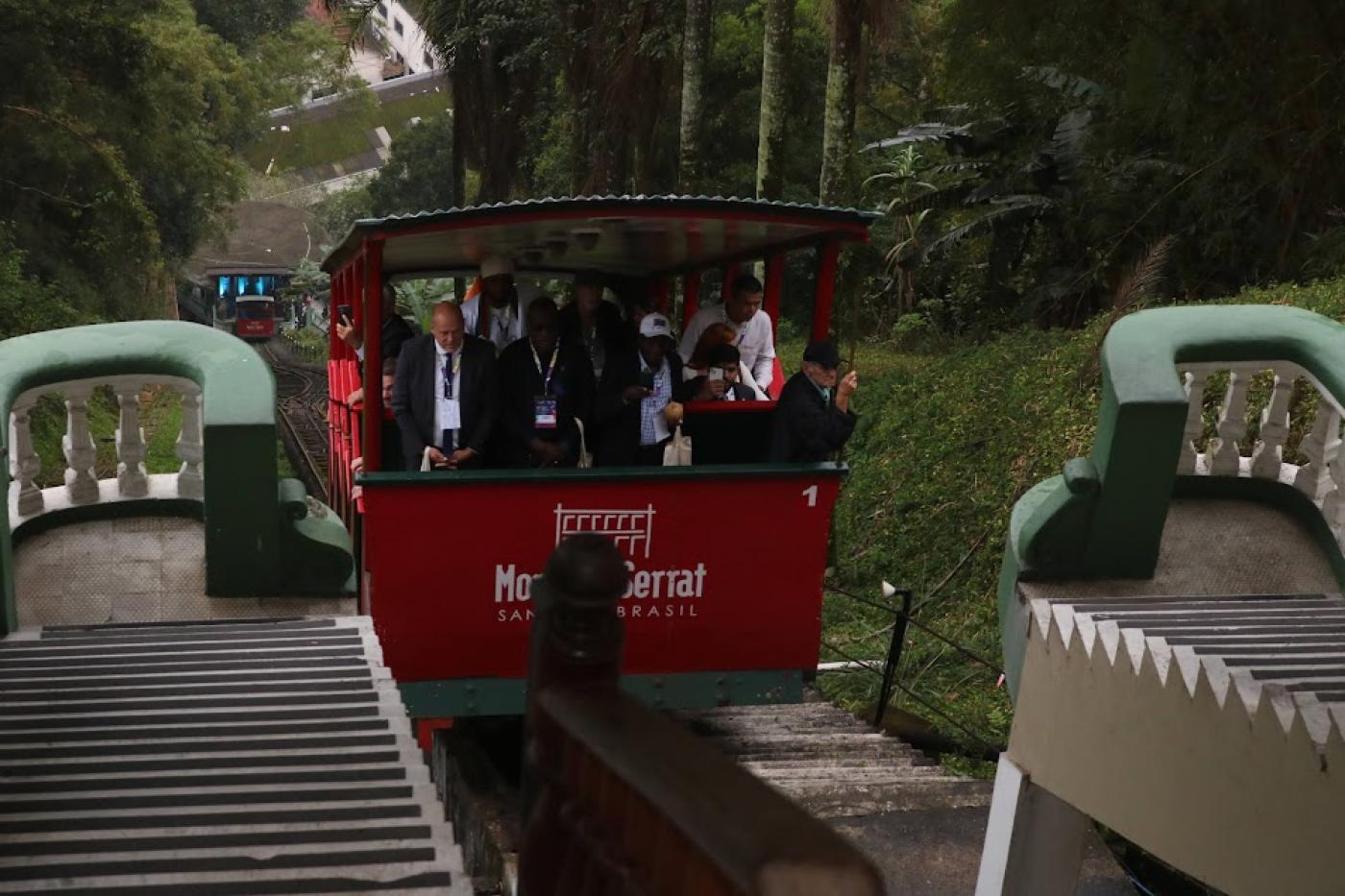 bondinho do monte serrat está em movimento de subida com vários passageiros. Há vegetação do morro ao fundo. #paratodosverem