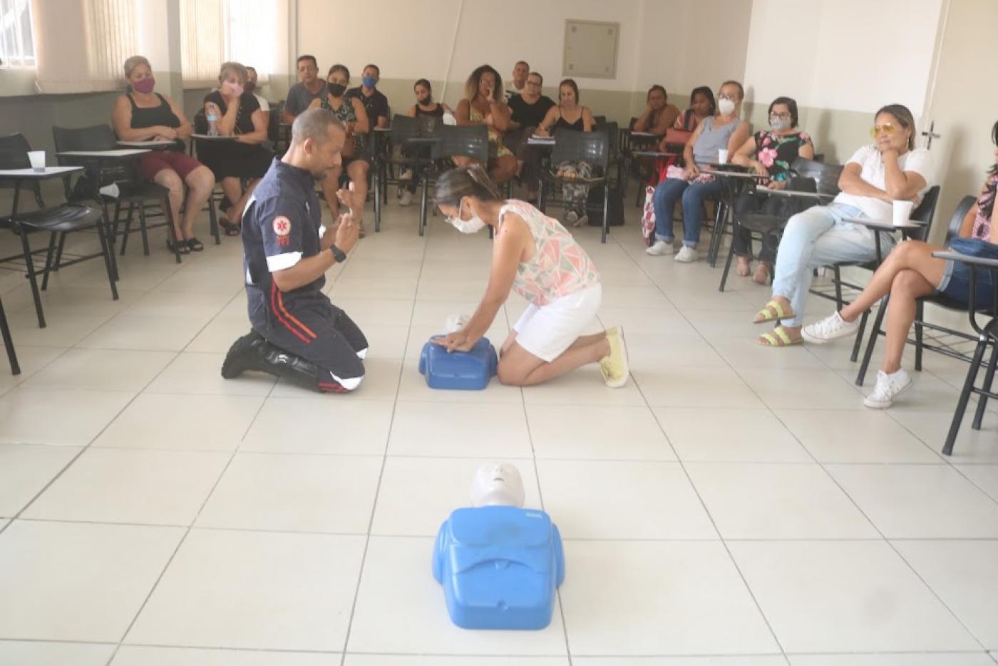 enfermeiro do Samu e uma mulher estão ajoalhados no chão. Entre eles, um boneco serve de simulação para primeiros socorros. Ela aperta o peito do boneco. Outras pessoas sentadas assistem. #paratodosverem