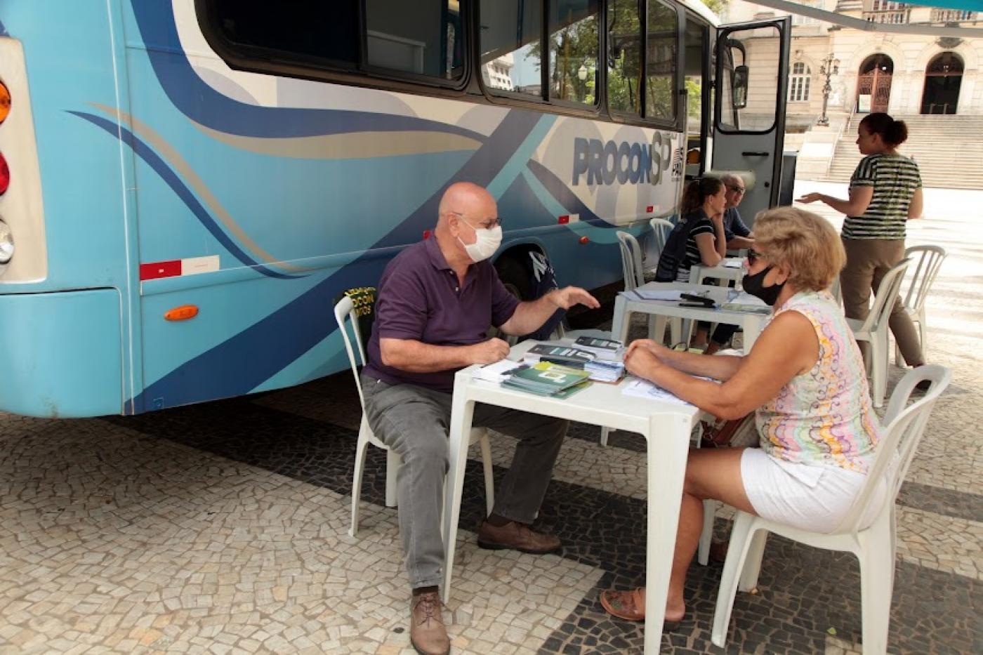 ônibus do procon parado na praça e um homem atendendo uma mulher, com ambos sentados à mesa. #paratodosverem