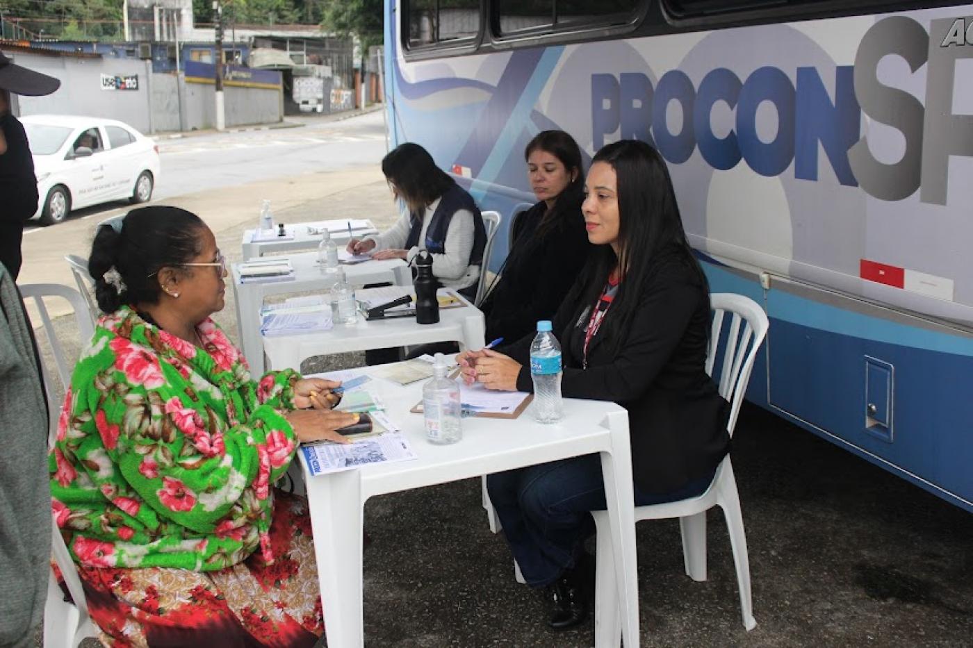 atendentes do procon estão sentadas à frente de van. Uma delas atende uma mulher. #paratodosverem