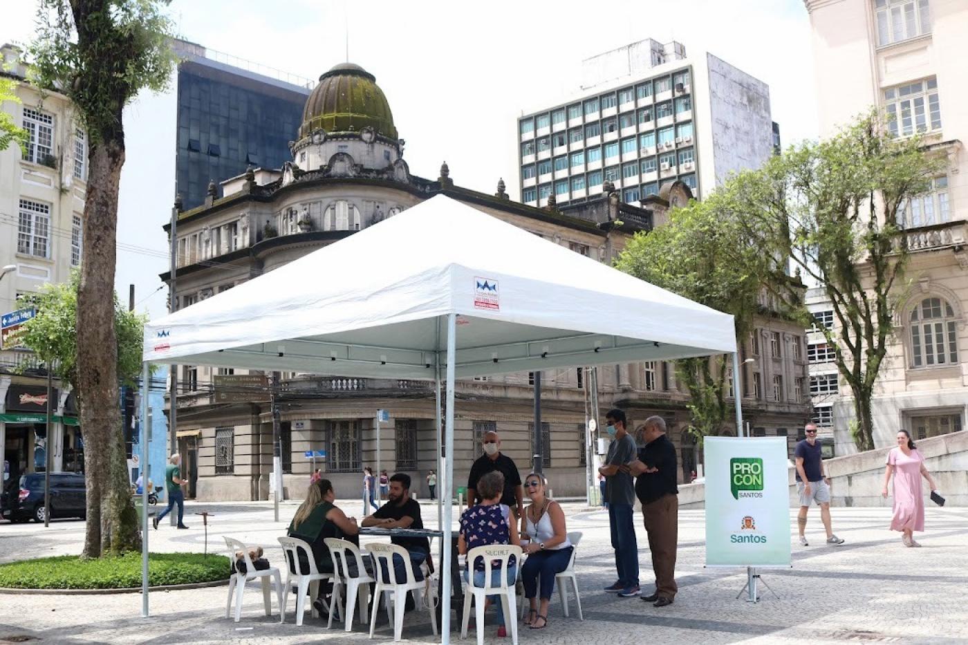 tenda armada em praça com banner do procon ao lado. Há pessoas sentadas. #paratodosverem