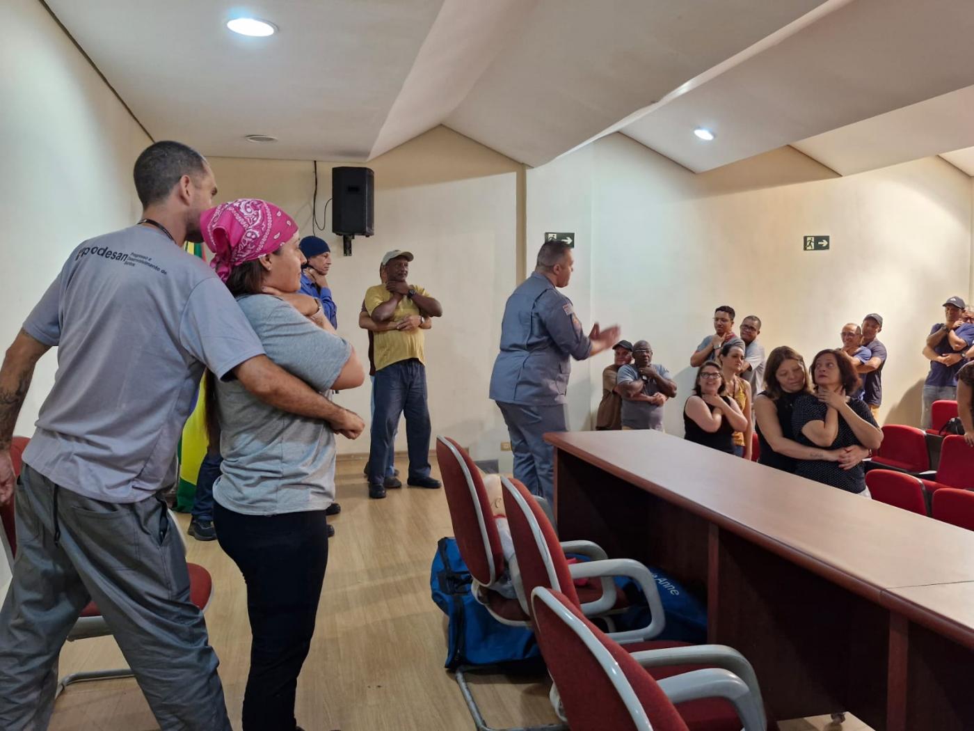 pessoas em pé em auditório simulando desengasgo de pessoa adulta. Um homem está orientando as duplas. #paratodosverem 