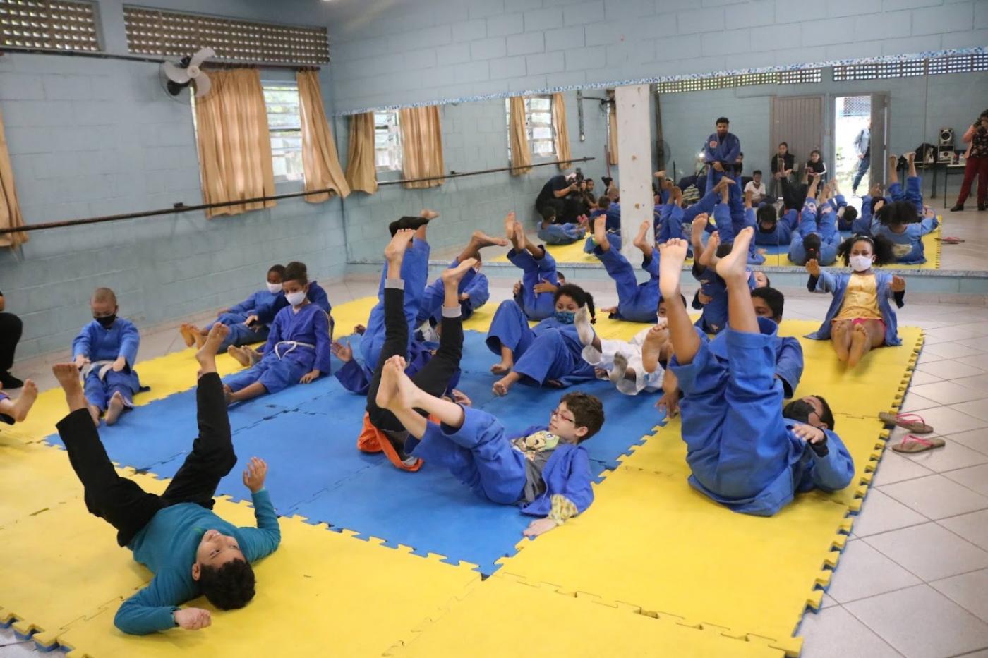 crianças vestidas com quimonos estão praticando judo sobre tatame. pelo movimento, elas parecem treinar queda no chão. #paratodosverem