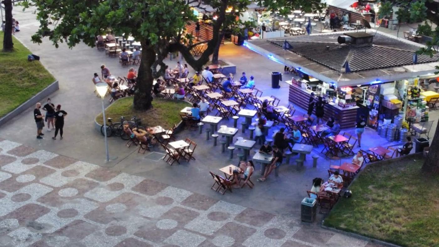 vista do alto, por drone, de quiosque no jardim da orla, com mesas em torno e público. #paratodosverem