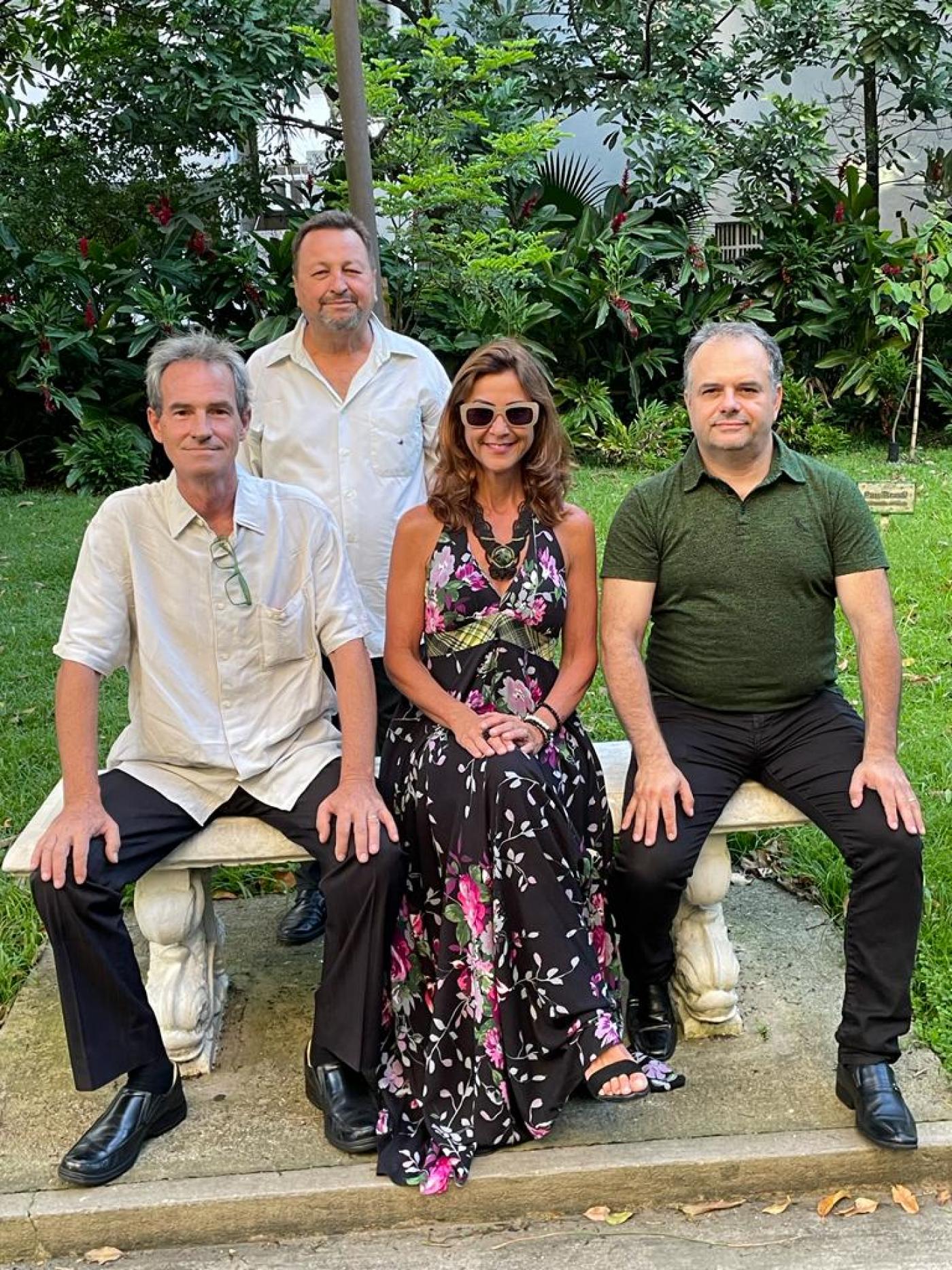 o quarteto posa para foto em banco de jardim. Dois homens e uma mulher estão sentados. Outro homem está em pé, atrás. #paratodosverem
