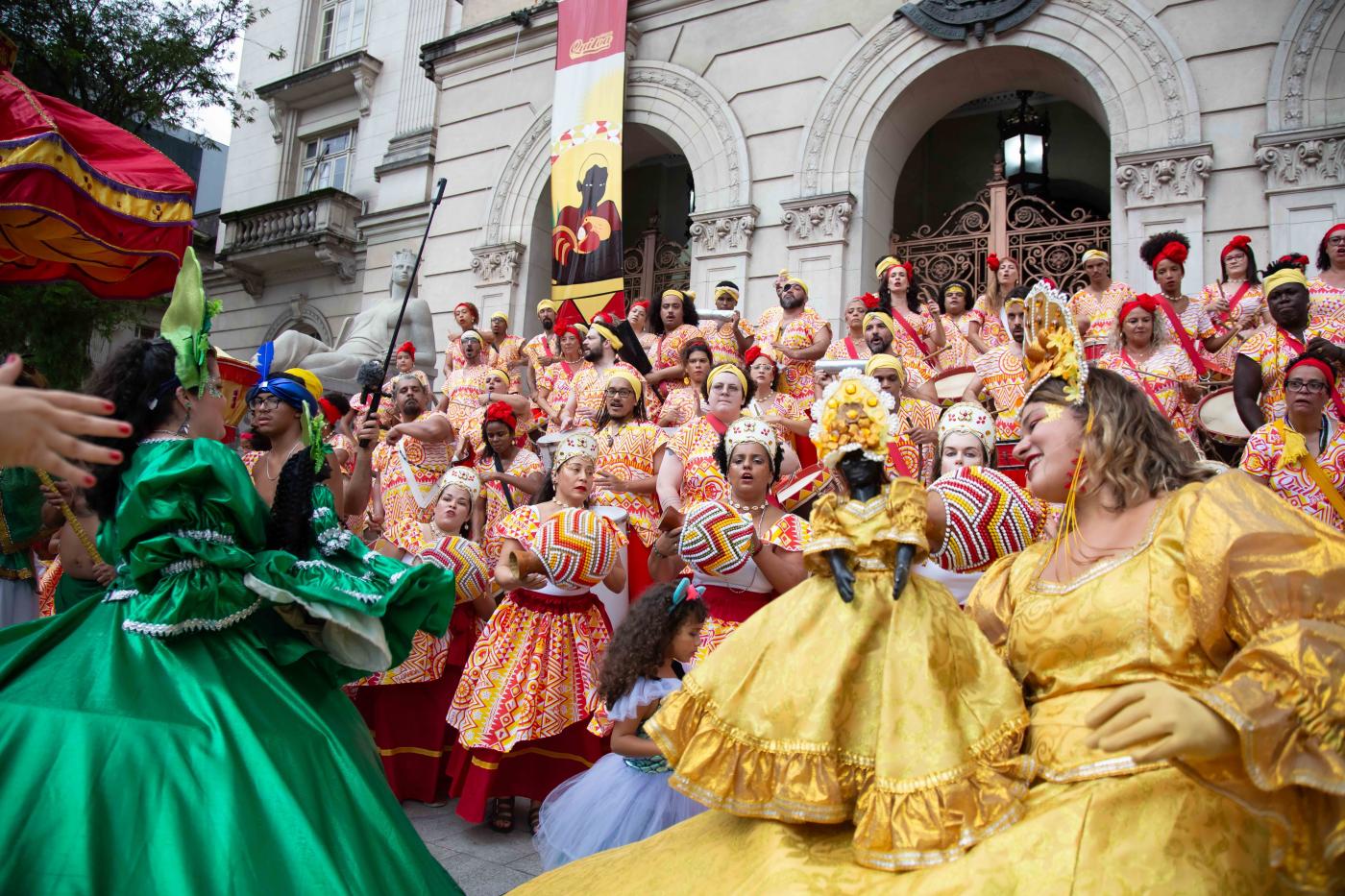 grupo quiloa fantasiado com roupas de desfile de carnaval coloridas dança na escadaria da prefeitura. #paratodosverem 