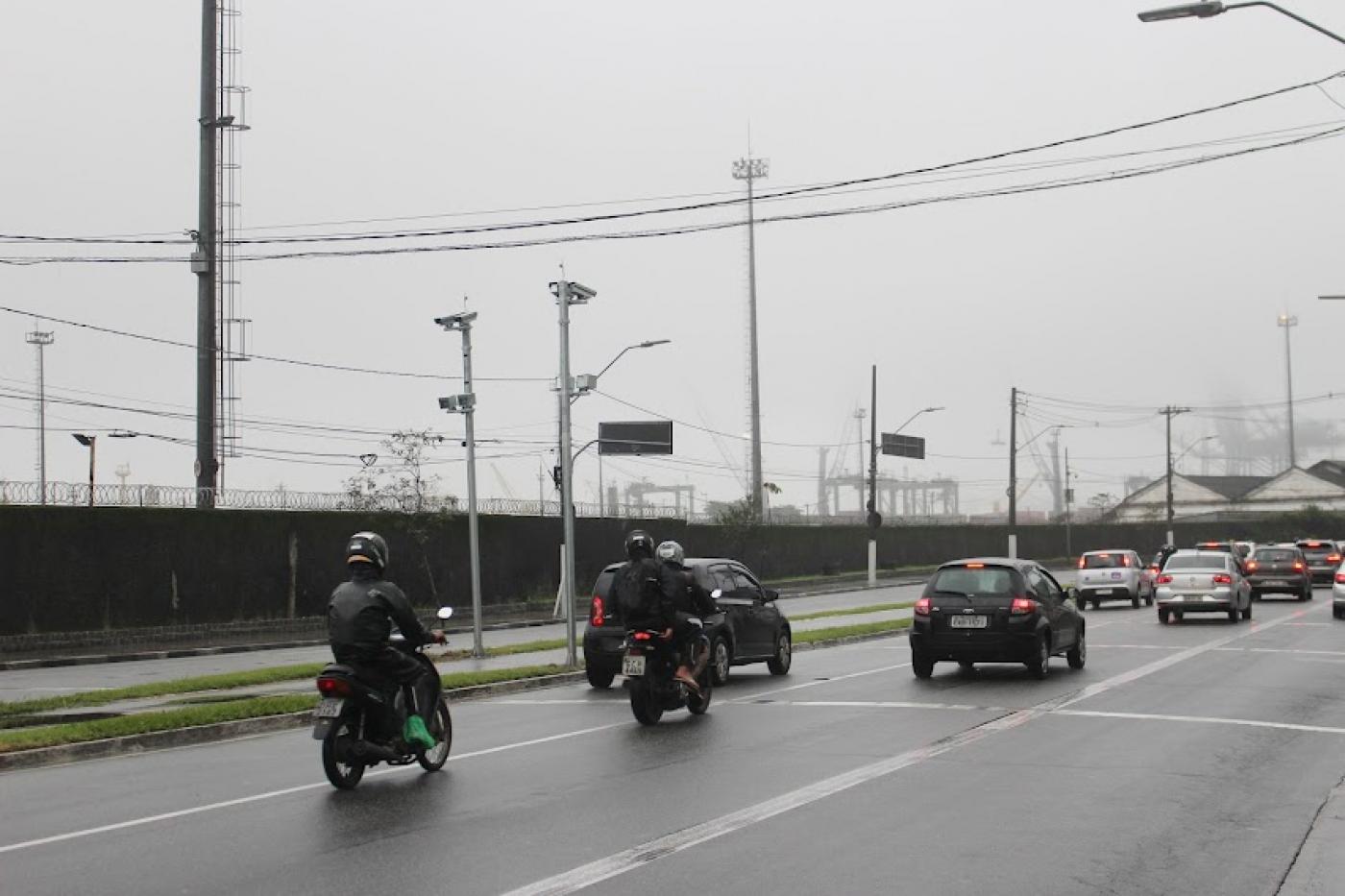 pista de avenida com motos e carros passando. Ao fundo, postes com radares no topo. #paratodosverem