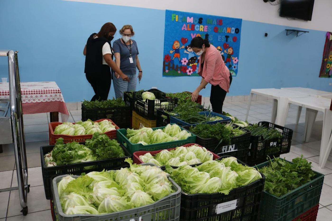 três mulheres observam caixas com alimentos em pátio de escola #paratodosverem