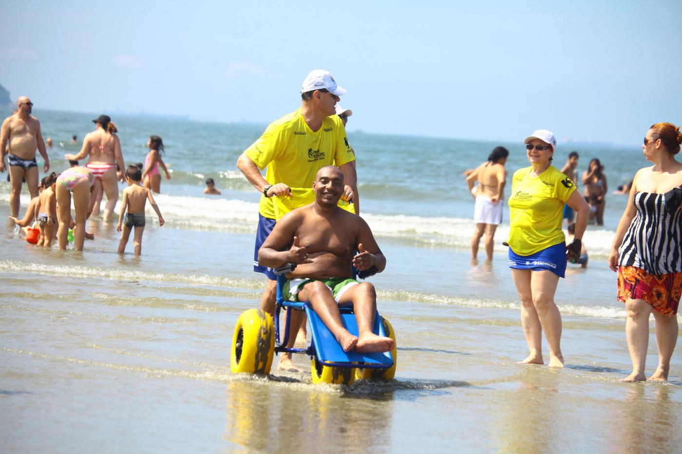 Homem está sentado em cadeira acessível ao mar. Outro homem dirige a cadeira. #Pracegover