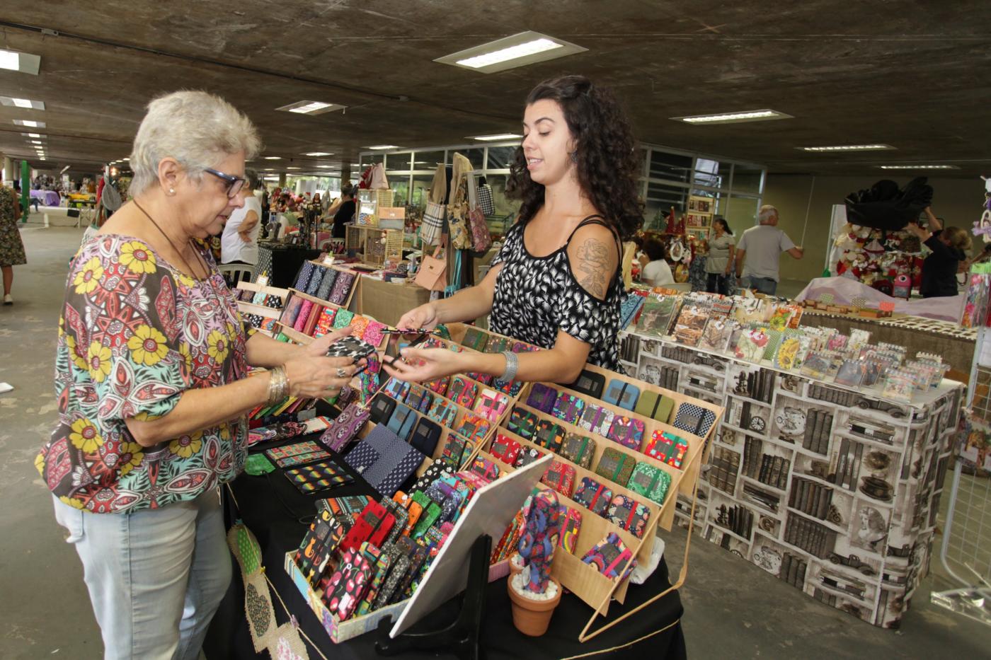 Duas  mulheres estão uma de frente para a outra. Entre elas há uma mesa com produtos artesanais em exposição. #Pracegover