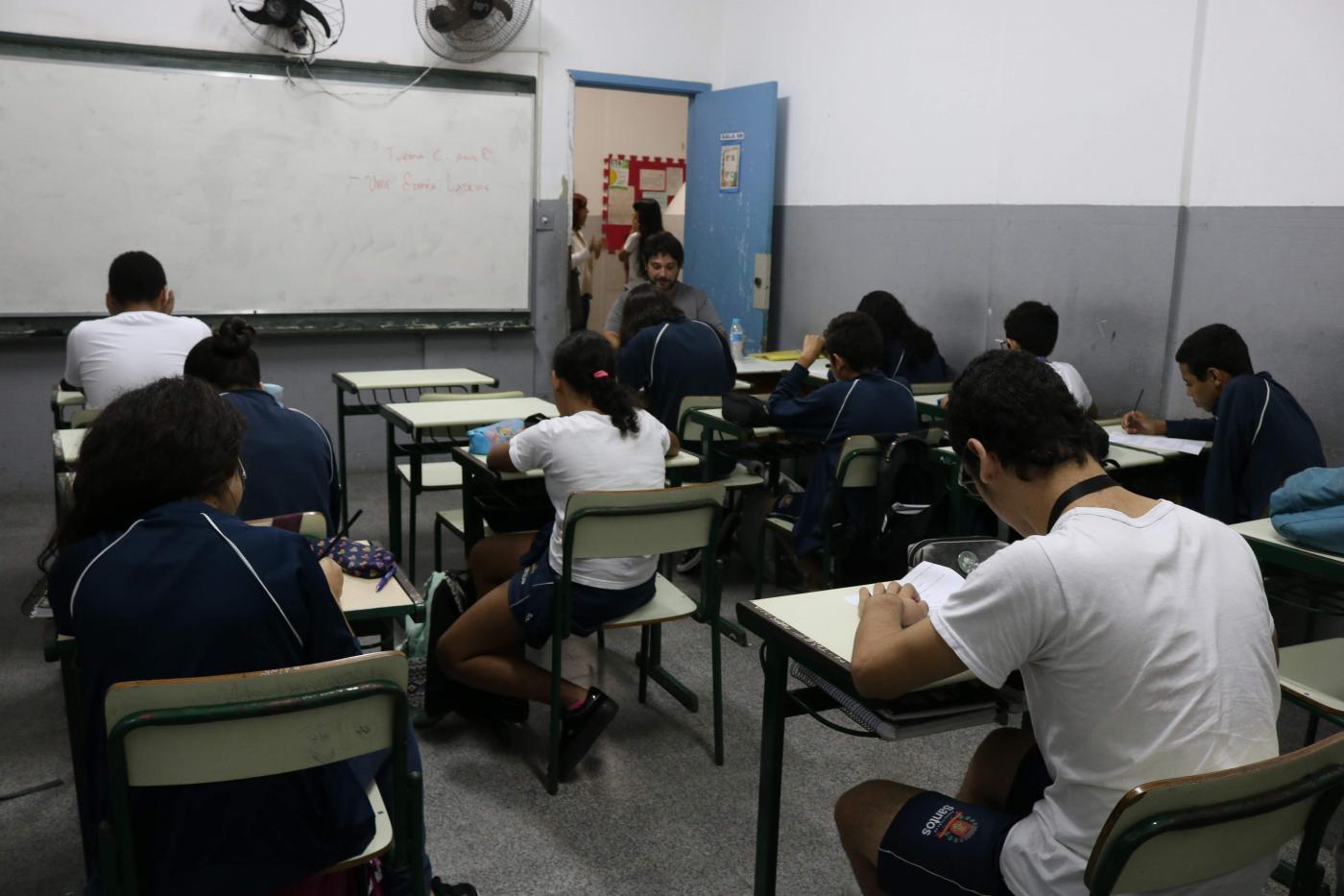 Alunos estão em sala de aula, sentados escrevendo em papéis. Todos estão de costas para a imagem. #Pracegover