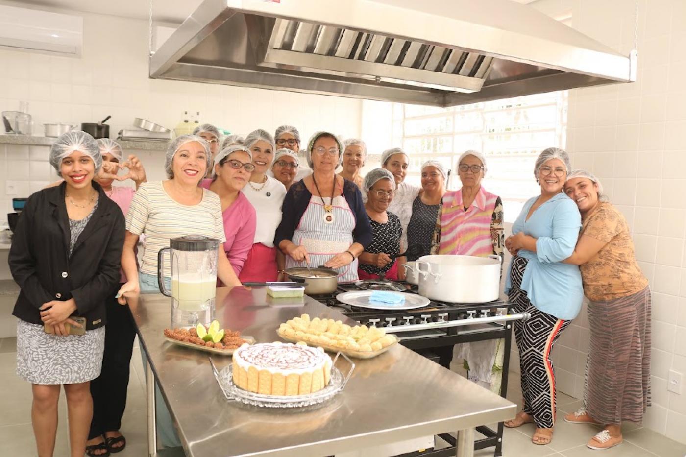 Grupo de mulheres à frente de mesa com doces e salgados. #paratodosverem