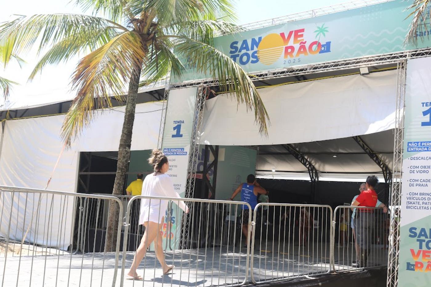 a fachada de uma tenda onde se lê no alto Santos Verão. Uma mulher está entrando no recinto. #paratodosverem 