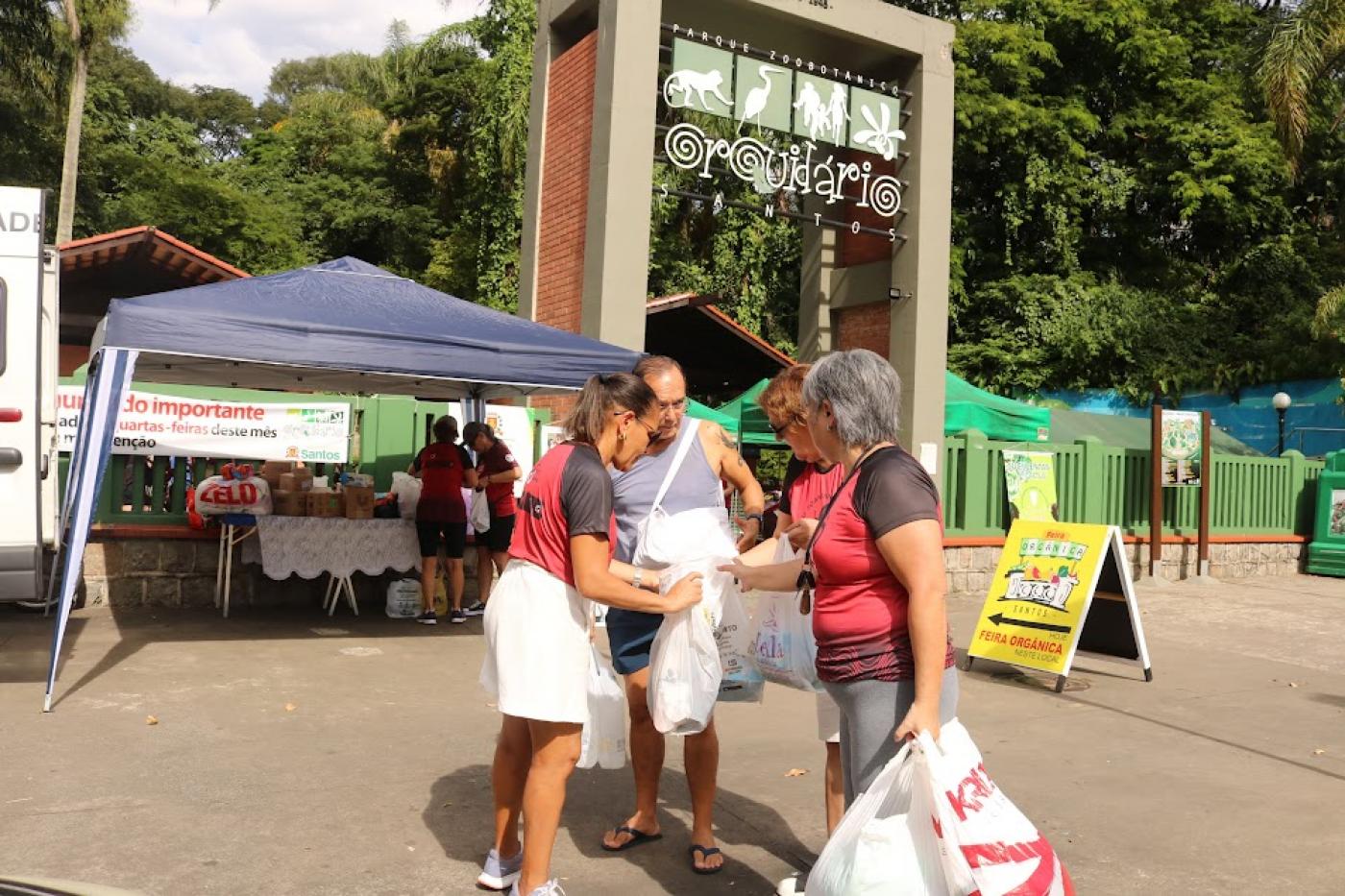 pessoas seguram sacos com doações na porta do orquidário. Ao fundo há uma tenda onde estão concentrados pacotes. #paratodosverem