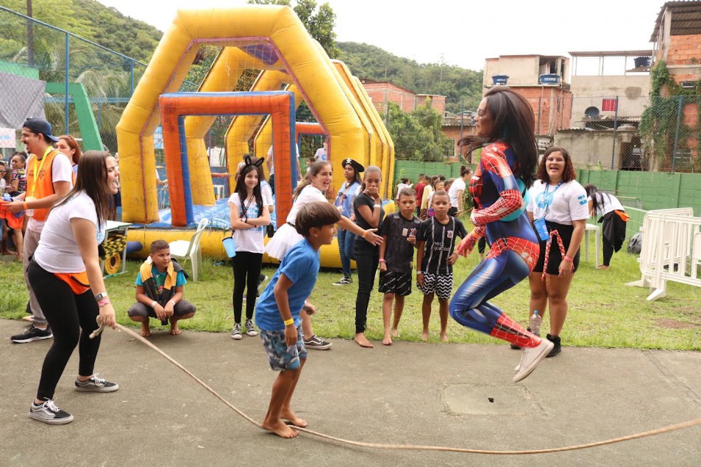 criança e mulher vestida de mulher maravilha pulam corda frente a frente. ao fundo uma estrutura de futsabão. Várias crianças no ambiente. #paratodosverem