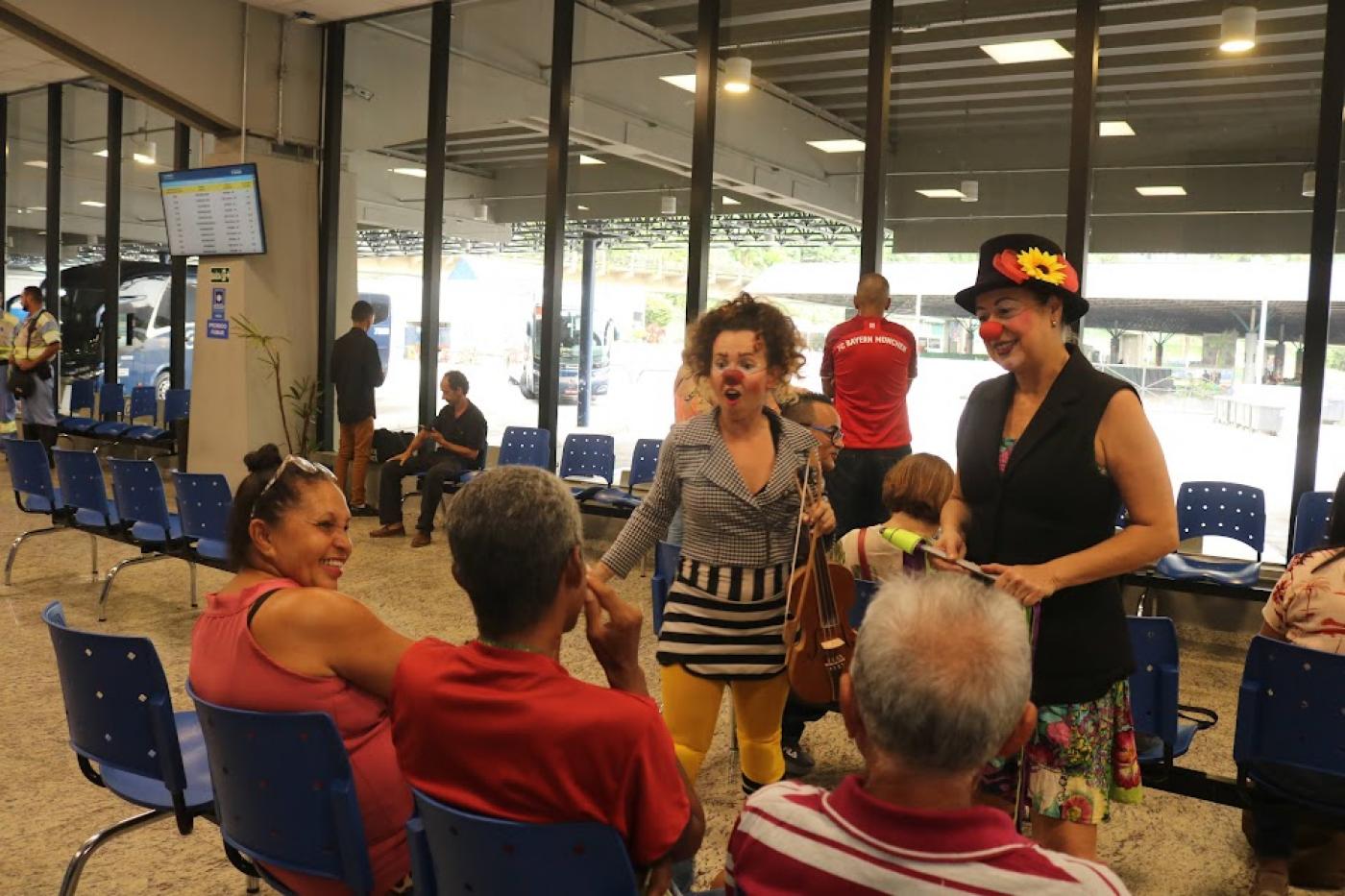 artistas vestidas de palhaça conversam com três pessoas que estão sentadas. Uma mulher ri. #paratodosverem