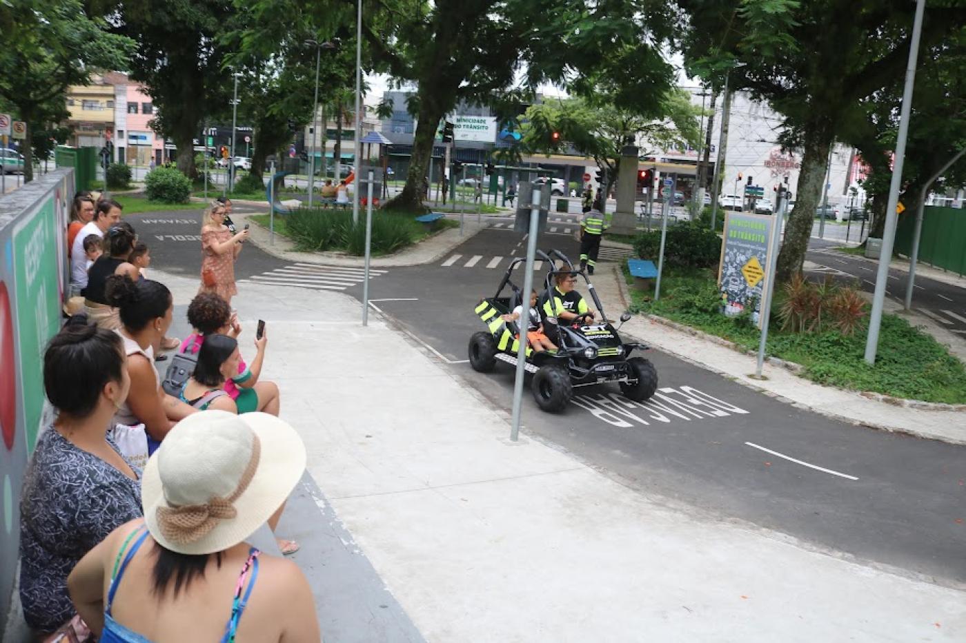 adultos próximos a um muro observam criança dentro de um minibug que passa na pista da praça. #paratodosverem 