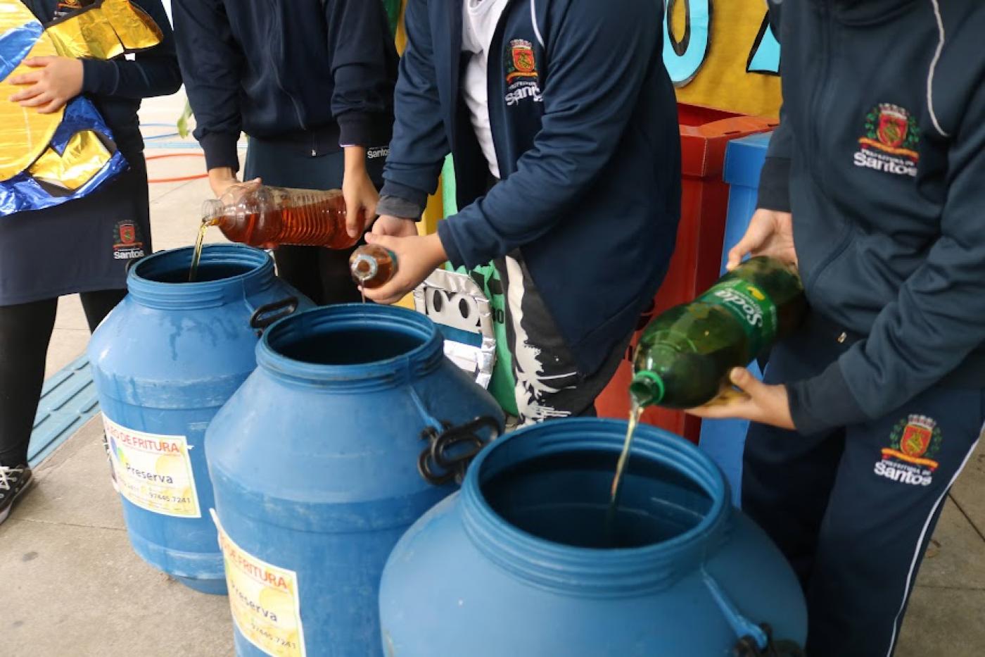 alunos uniformizados despejam óleo de garrafas pet para três galões. #paratodosverem