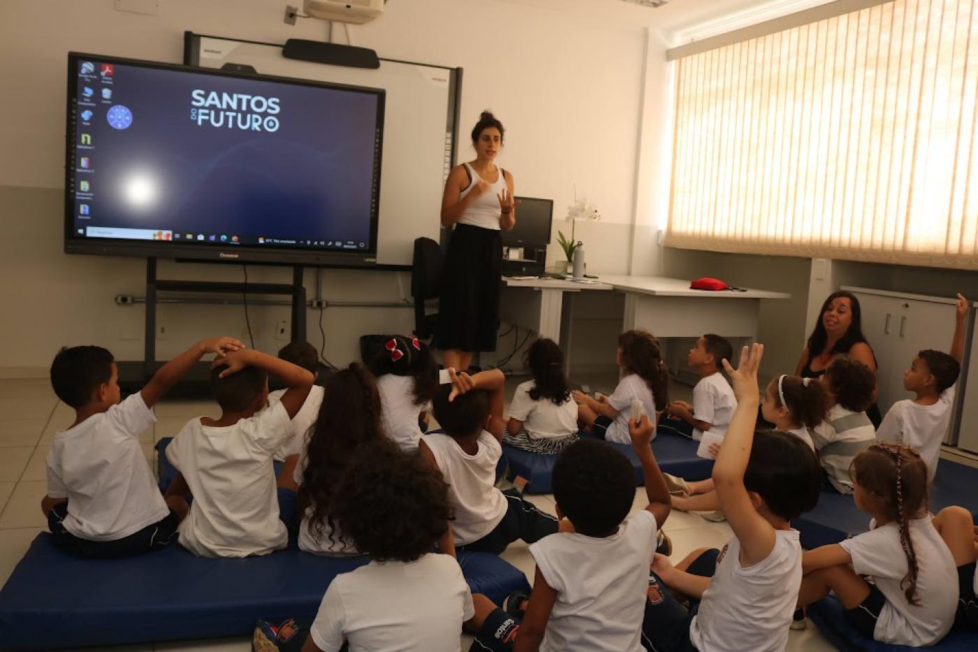 CRIANÇAS ESTÃO SENTADAS EM TATAMES NO CHÃO, algumas com os braços levantados para responder à professora que está à frente de todas. Há um telão ao fundo. #paratodosvere 