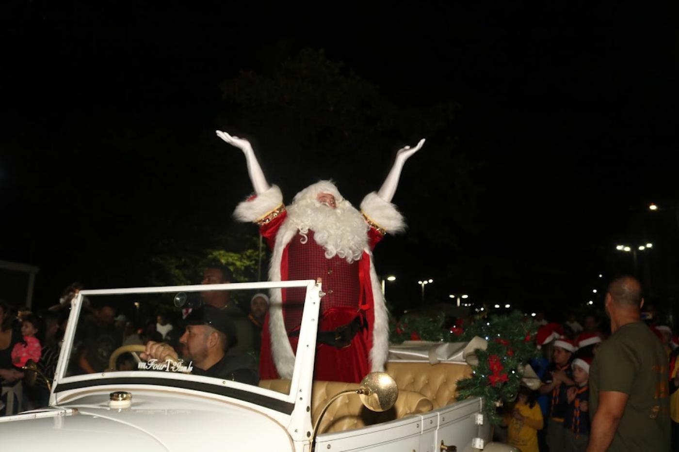 papai noel ergue os braços dentro de um carro aberto cumprimentado o público. O carro é dirigido por um motorista. É noite. #paratodosverem 