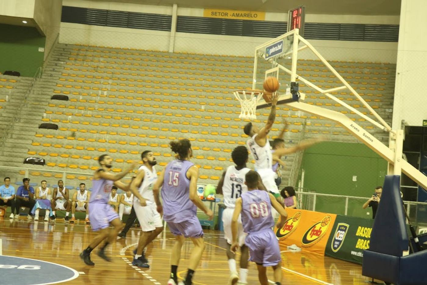 jogo de basquete. Um jogador salta com a bola em direção à cesta, bem próximo à ela. Outros jogadores estão em volta. #paratodosverem