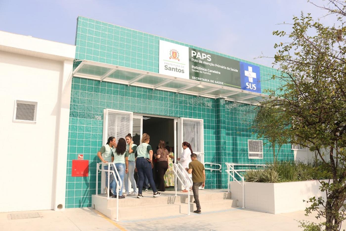 fachada de policlínica com pessoas na pequena escada de alvenaria que dá acesso ao prédio. #paratodosverem 