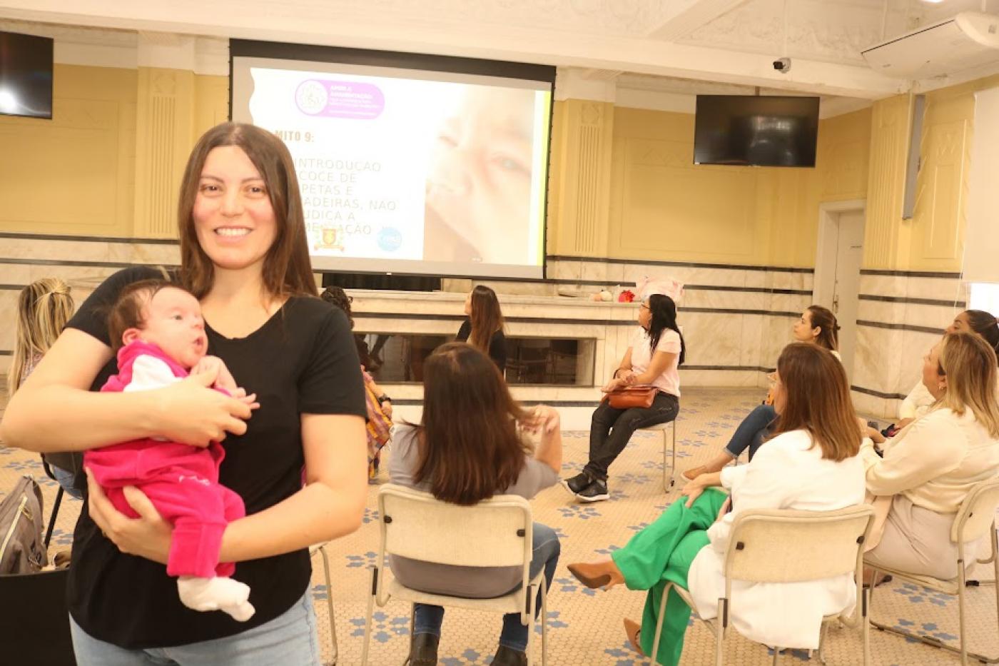 em primeiro plano, mãe segura bebê no colo em pé e sorrindo para a a foto. Ao fundo, uma roda com mulheres conversando. #paratodosverem 