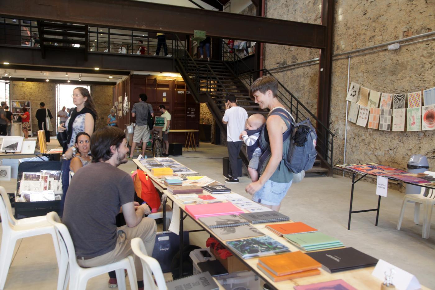 Livros expostos sobre uma bancada. Há um vendedor do lado esquerdo da foto. À direita, um homem olha as publicações