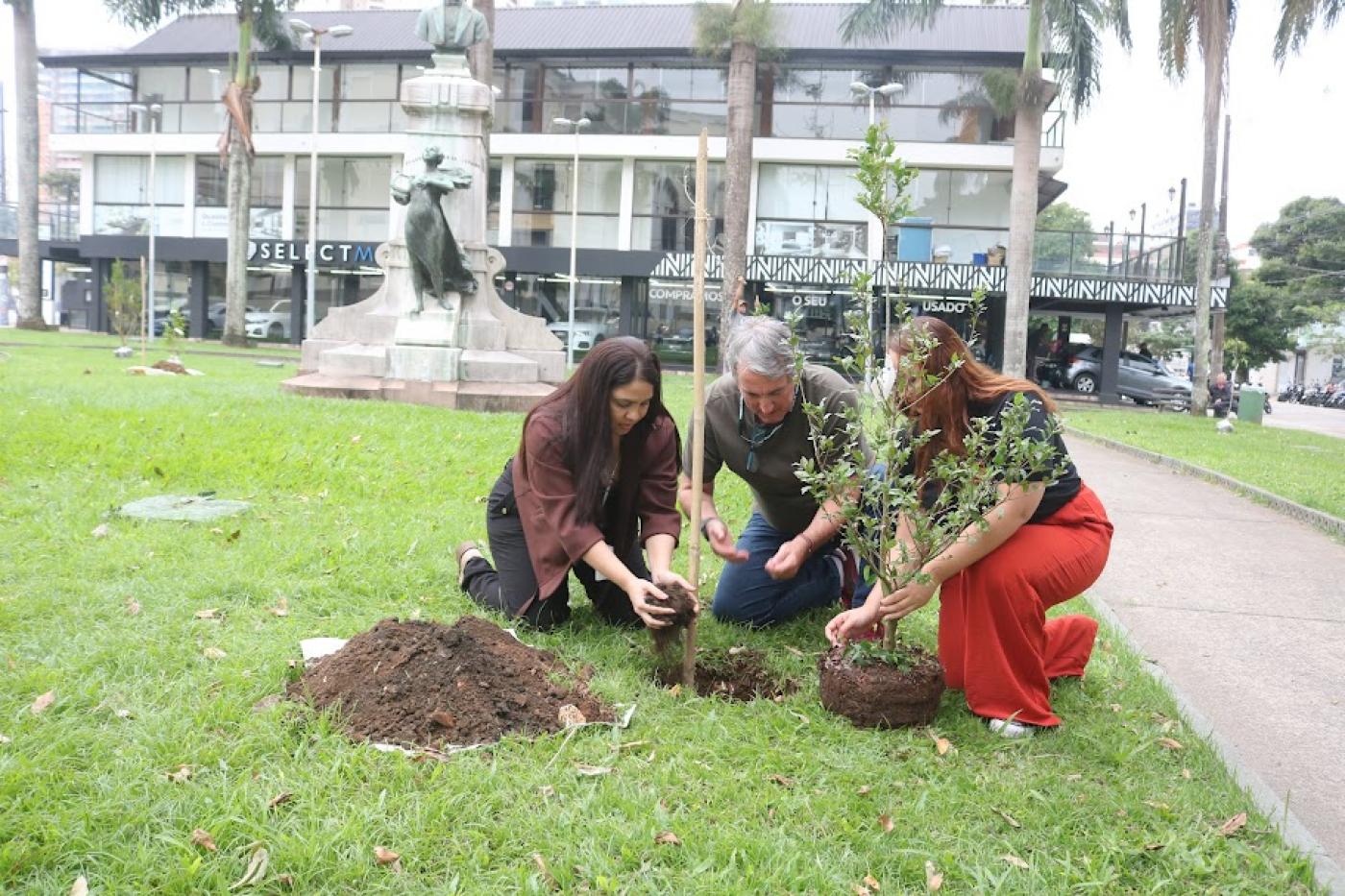 três pessoas plantam uma árvore em praça. #paratodosverem