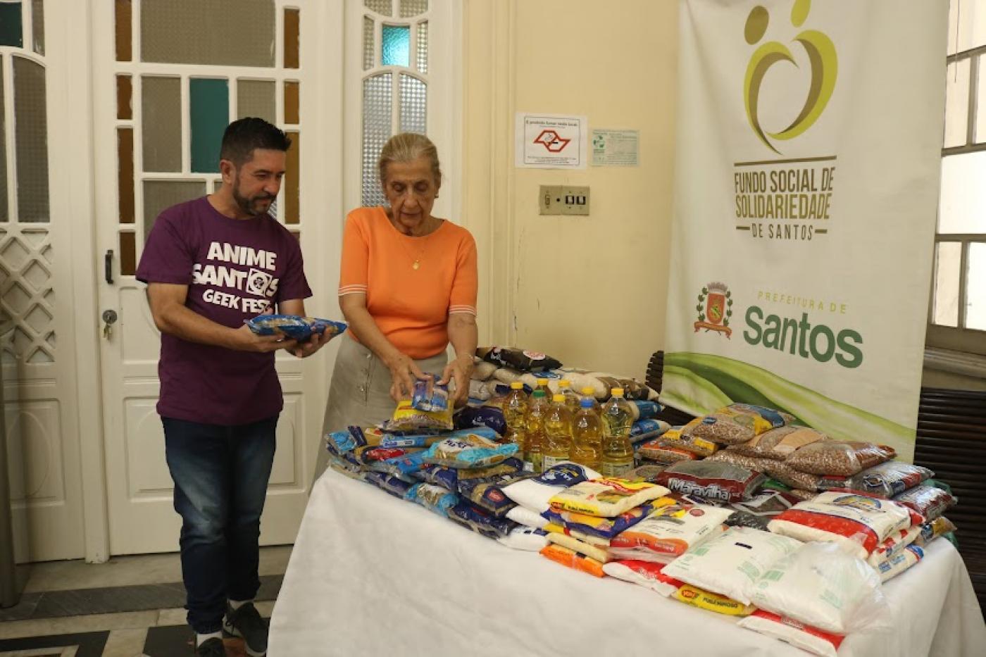 representante do evento e presidente do fundo social estão ao lado de mesa cheia de mantimentos. #paratodosverem