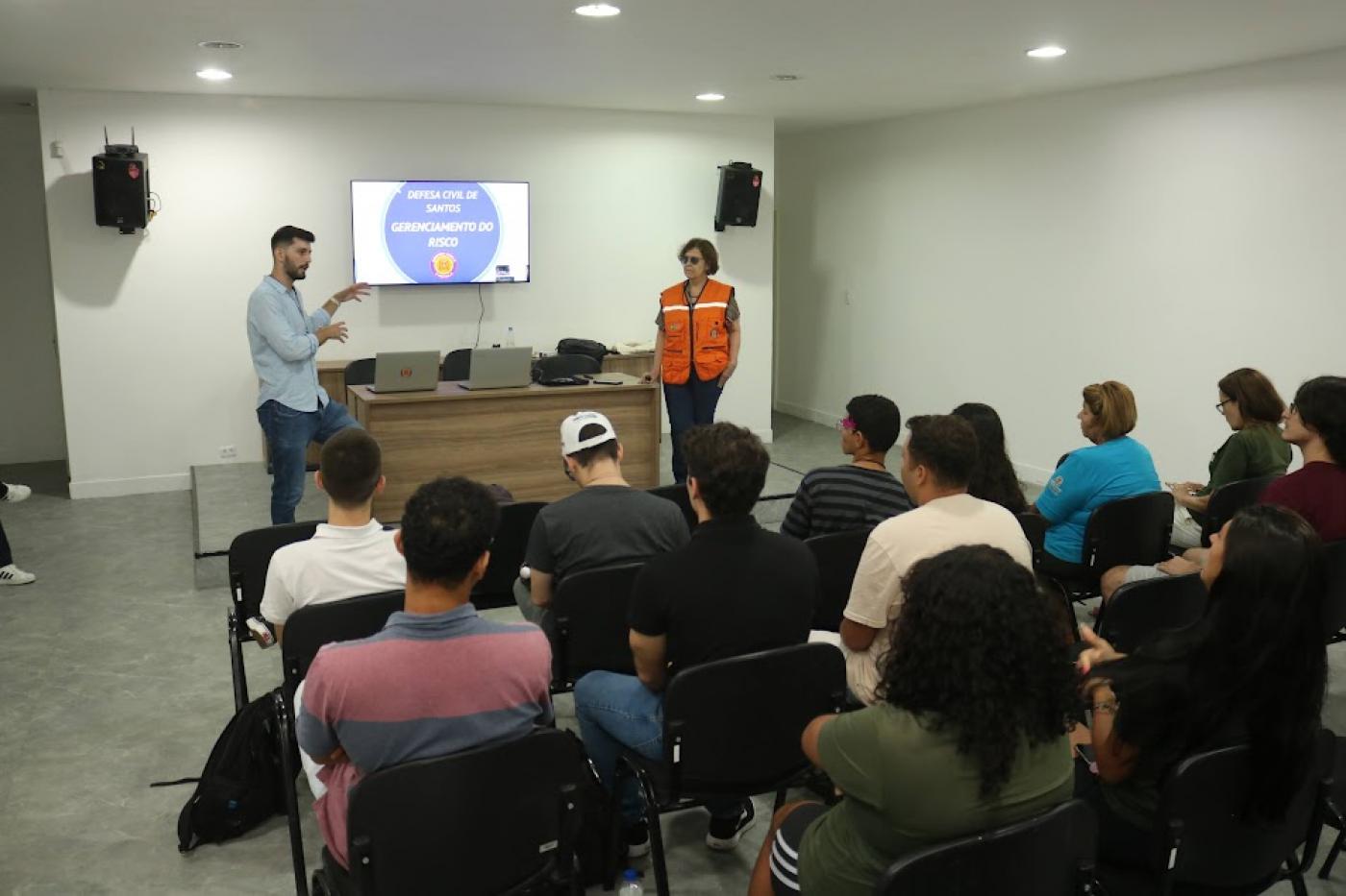 auditório com estudantes sentados e duas pessoas apresentando palestra. #paratodosverem