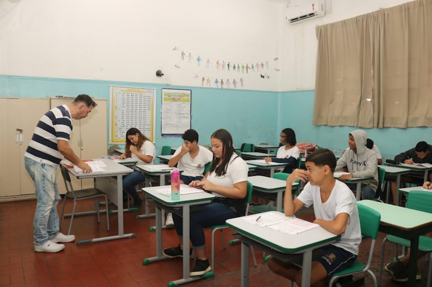 professor está em pé em sala diante de alunos sentados. #paratodoverem 