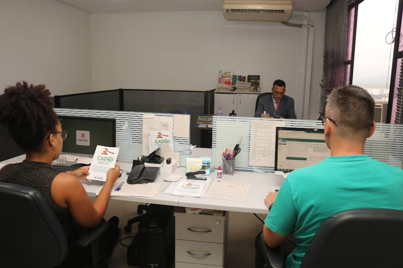 atendentens de costas para foto estão diante de computadores. A mulher, à esquerda, segura um folheto do cadoj. Ao fundo há um homem sentado a uma mesa.#paratodosverem 