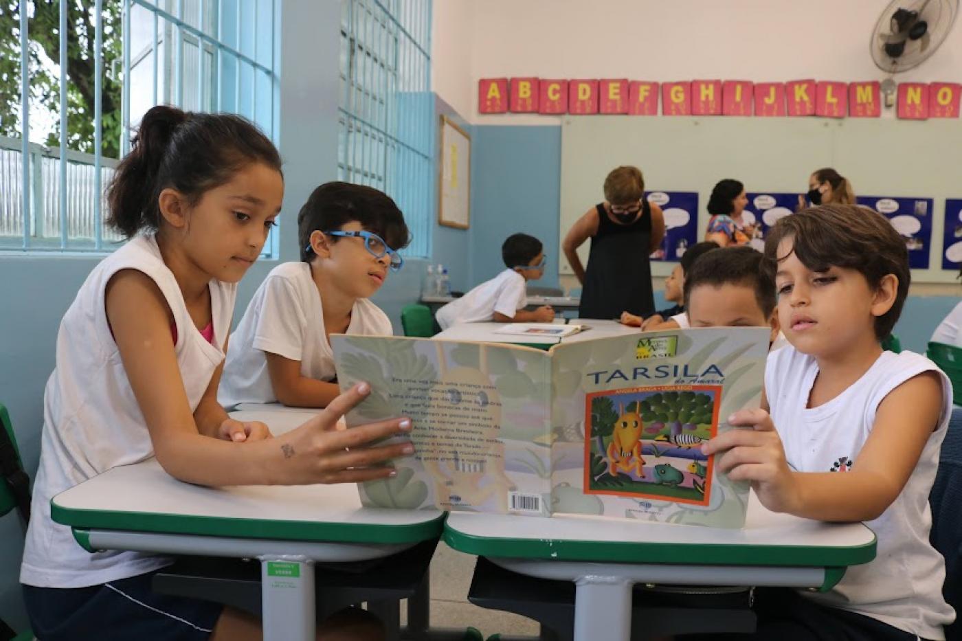 quatro crianças estão sentadas em torno de uma mesa. À frente, duas delas seguram um livro aberto, cada uma com a mão em uma das capas. #paratodosverem