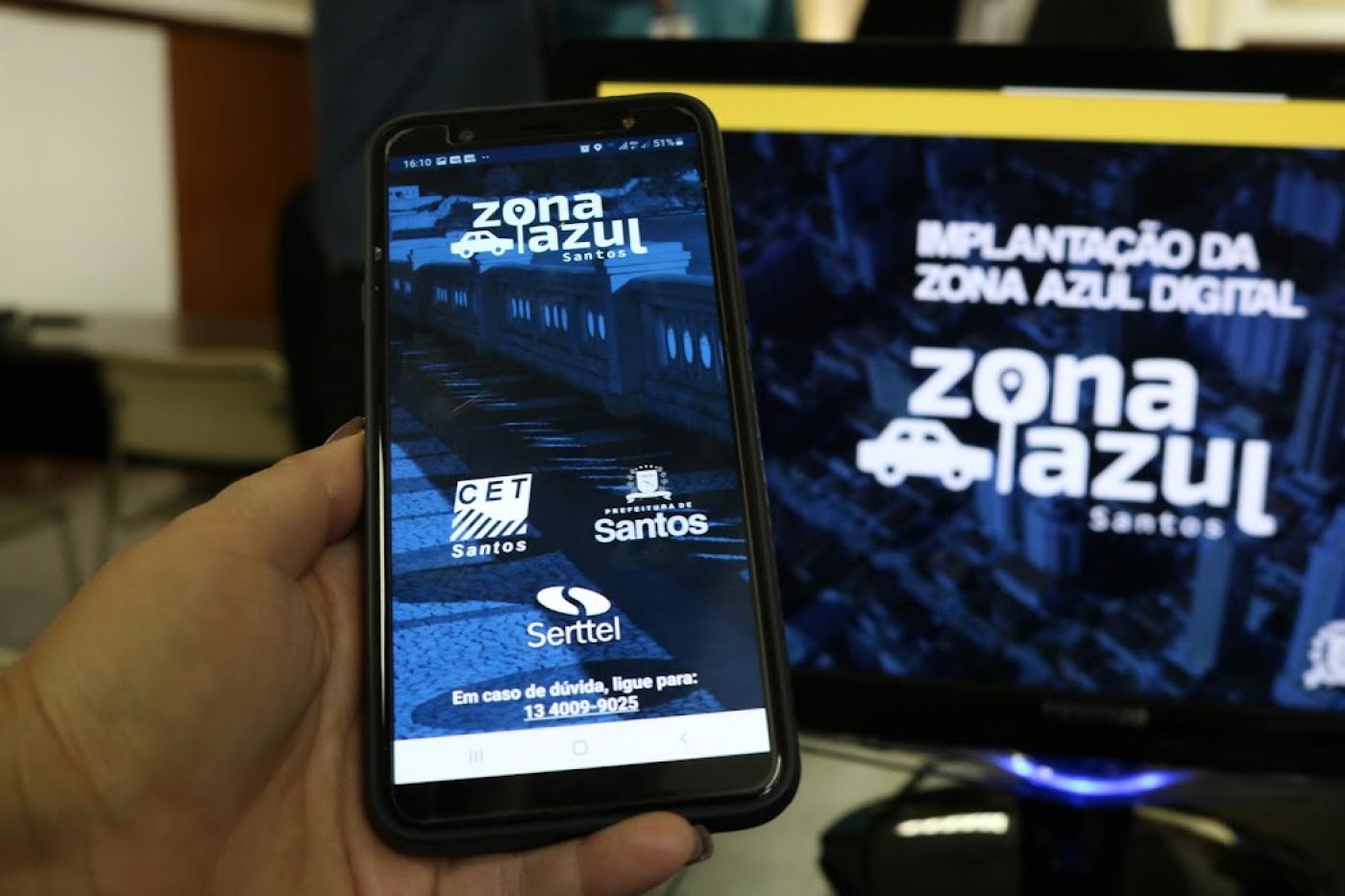 mão segurando celular com aplicativo zona azul aparecendo e tela com computador ao fundo #paratodosverem