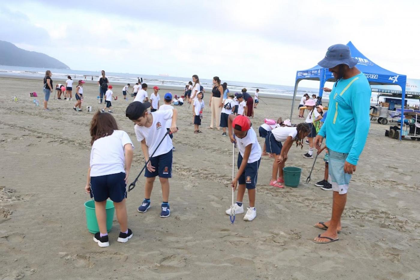 alunos uniformizados recolhem resíduos da areia da praia. #paratodosverem