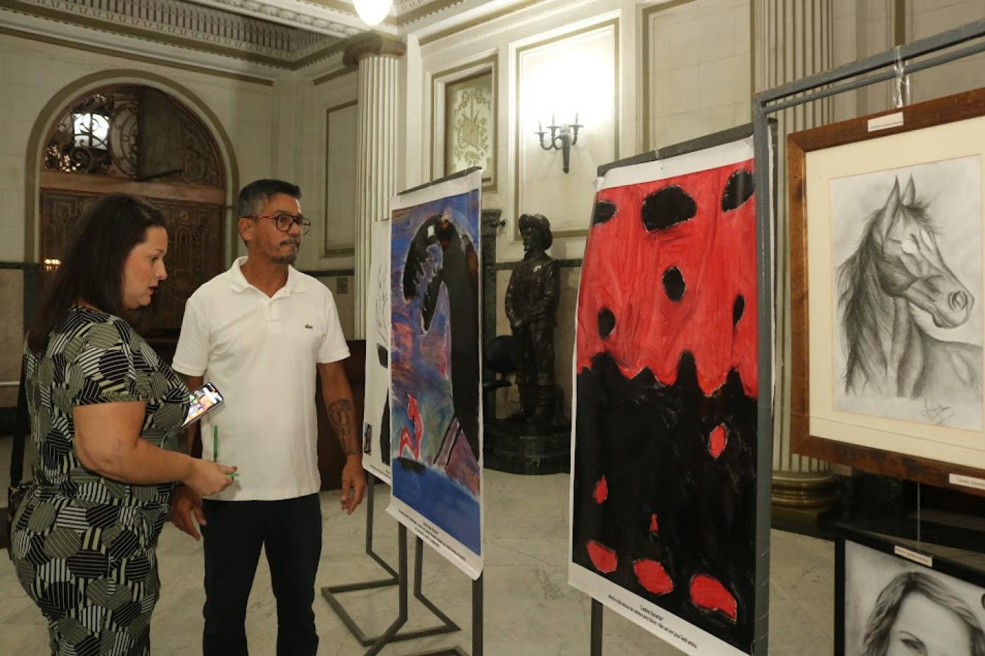 paineis com pinturas expostos em um salão com um homem e uma mulher olhando. #paratodosverem