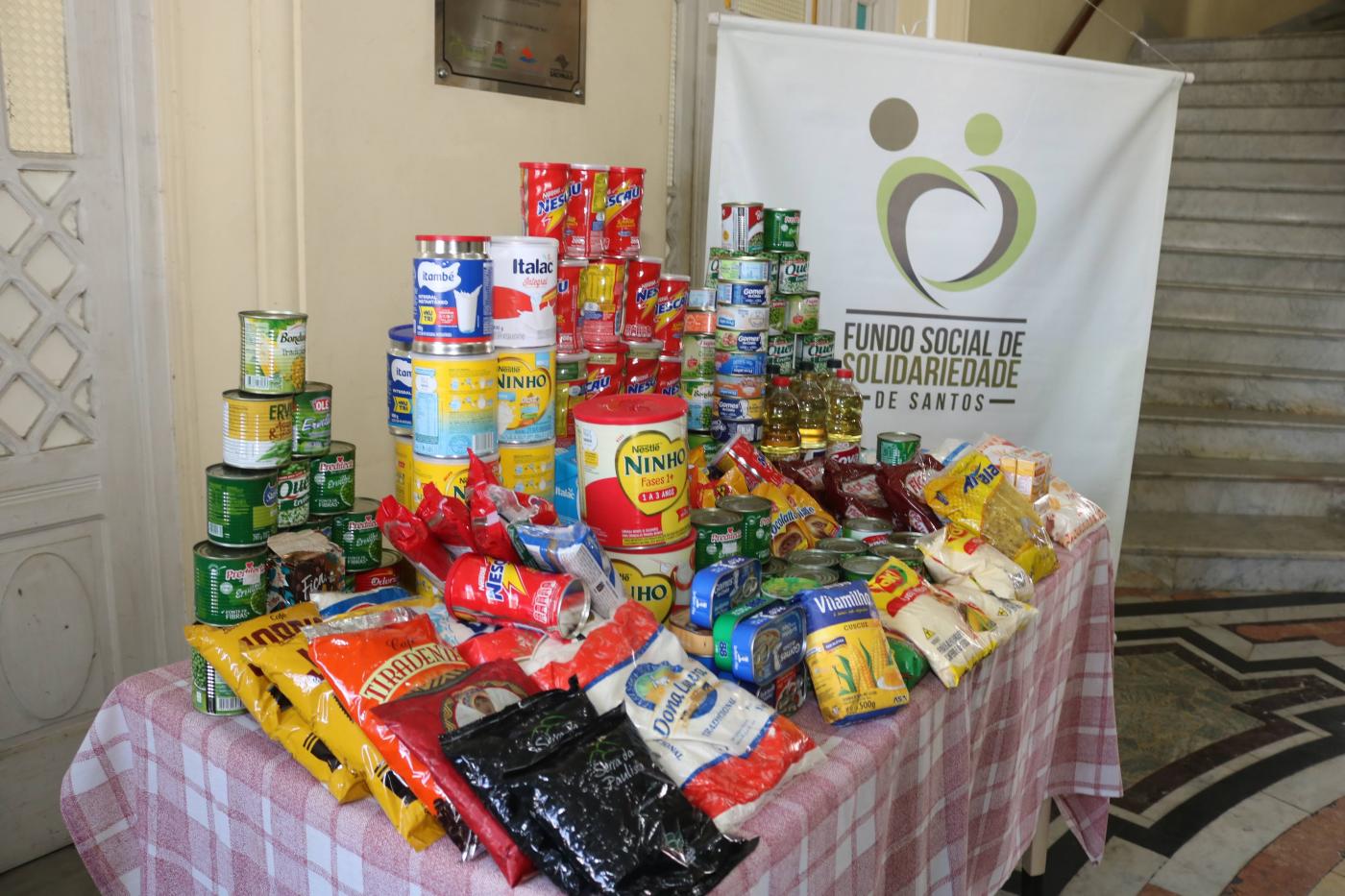 em uma mesa sacos de arroz, pacotes de feijão, latas de diversos produtos #paratodosverem