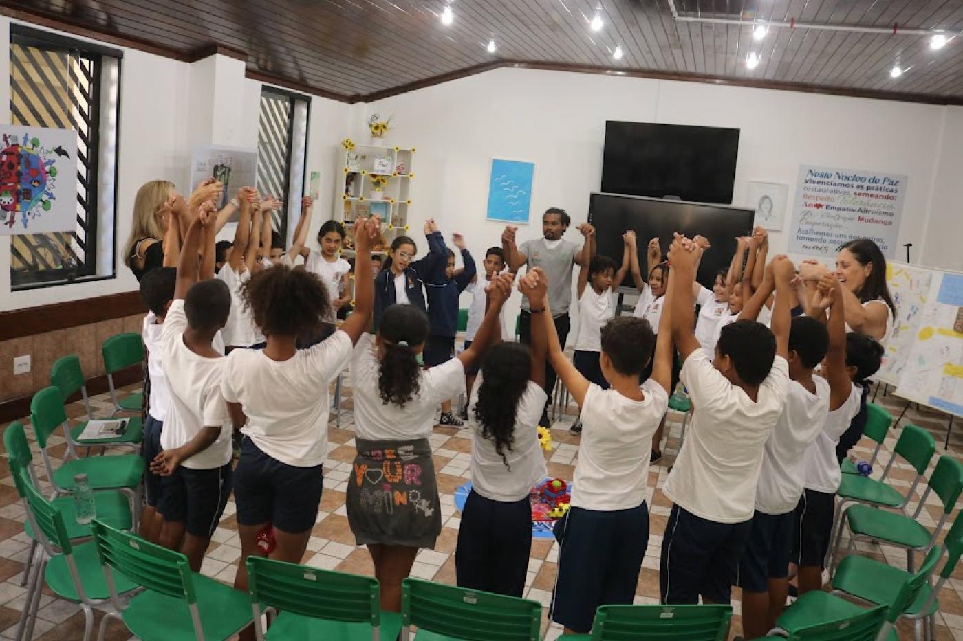 alunos estão de mãos dadas para o alto, reunidos em roda. #paratodosverem 
