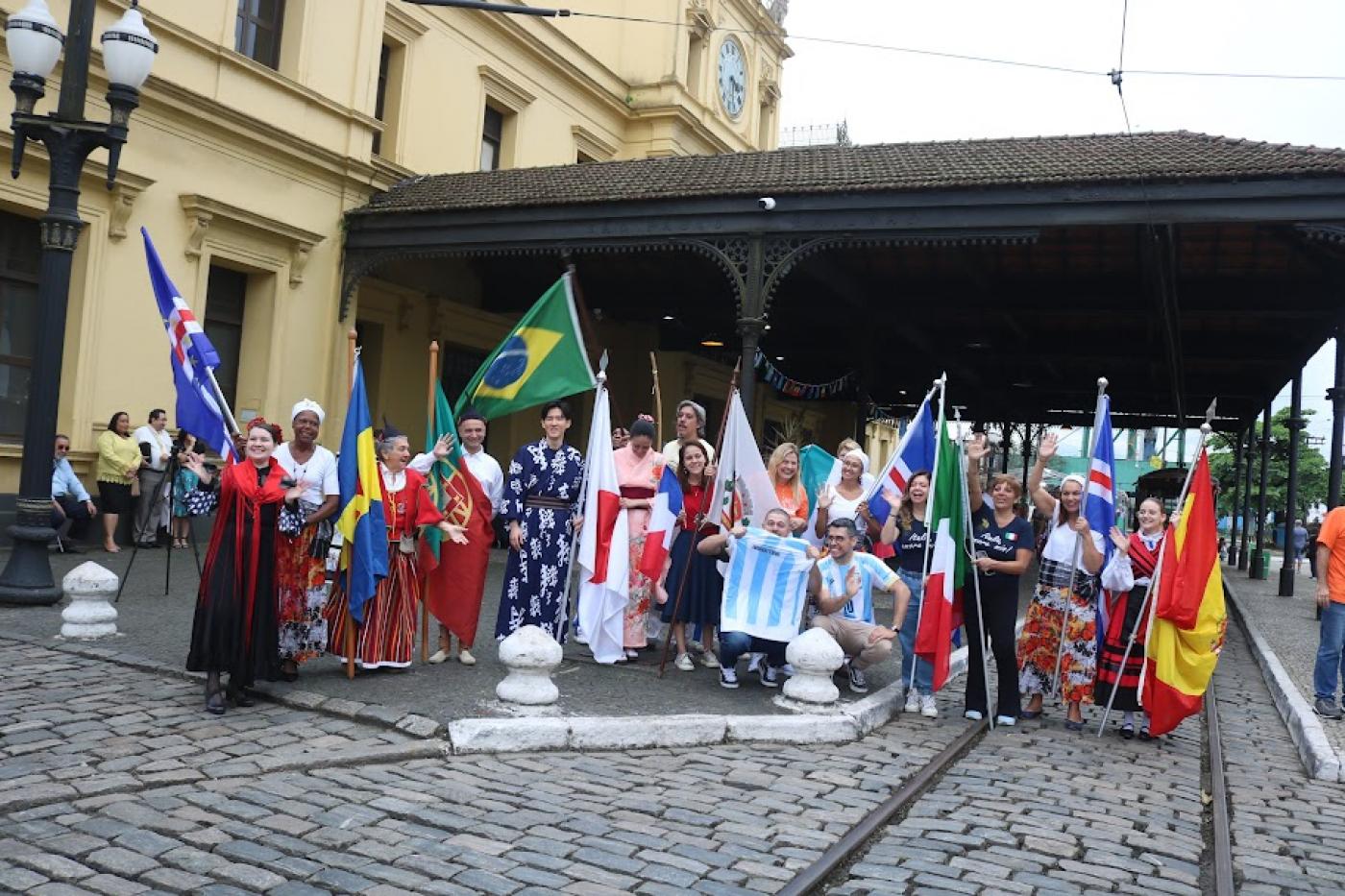 várias pessoas com trajes típicos e bandeiras de seus países posam para foto diante da estação do valongo. #paratodosverem