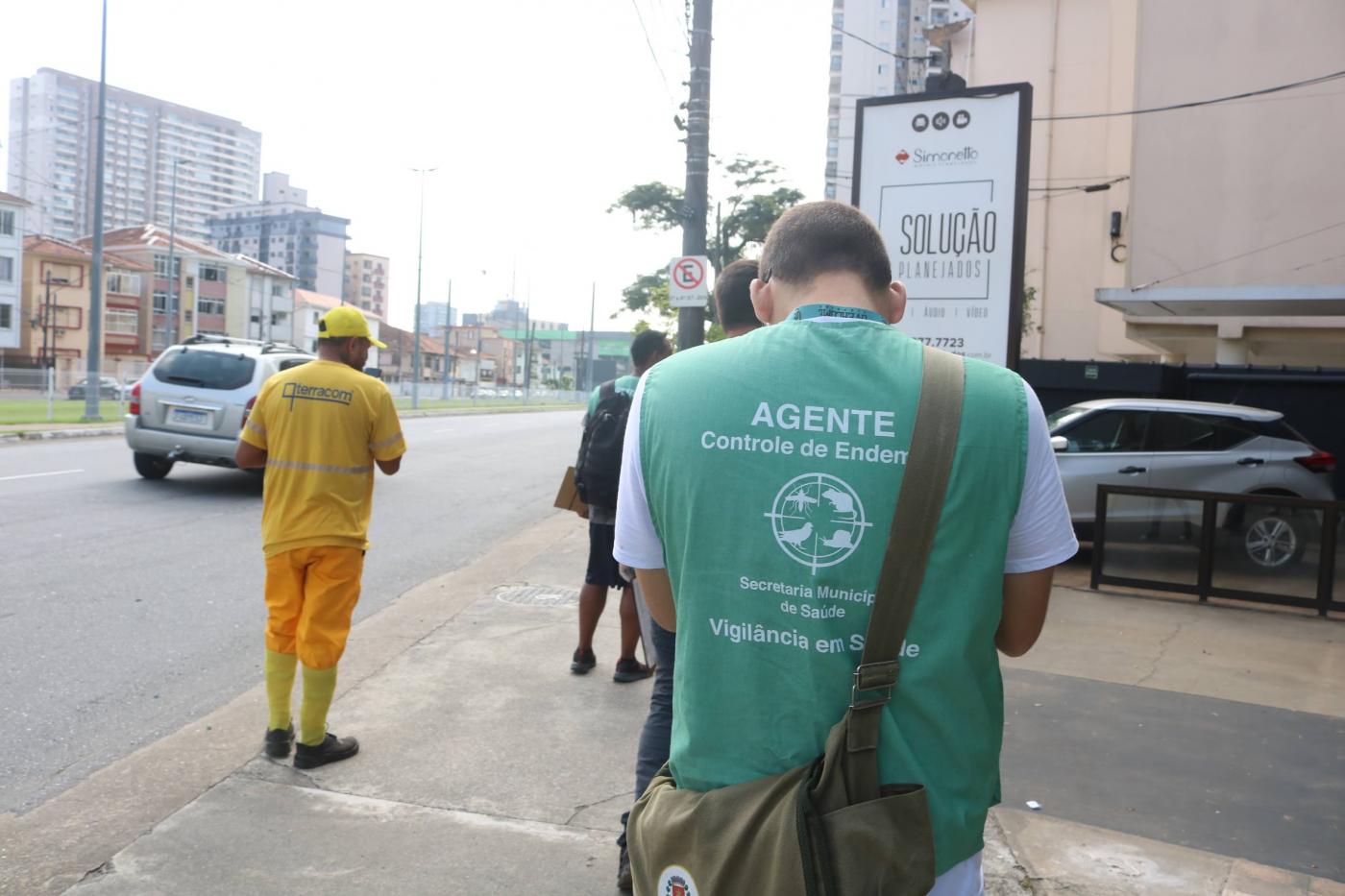 agente de combate à dengue caminha em rua, de costas para a foto, com evidência no colete verde. Ao lado esquerdo, um funcionário da terracom. #paratodosverem 