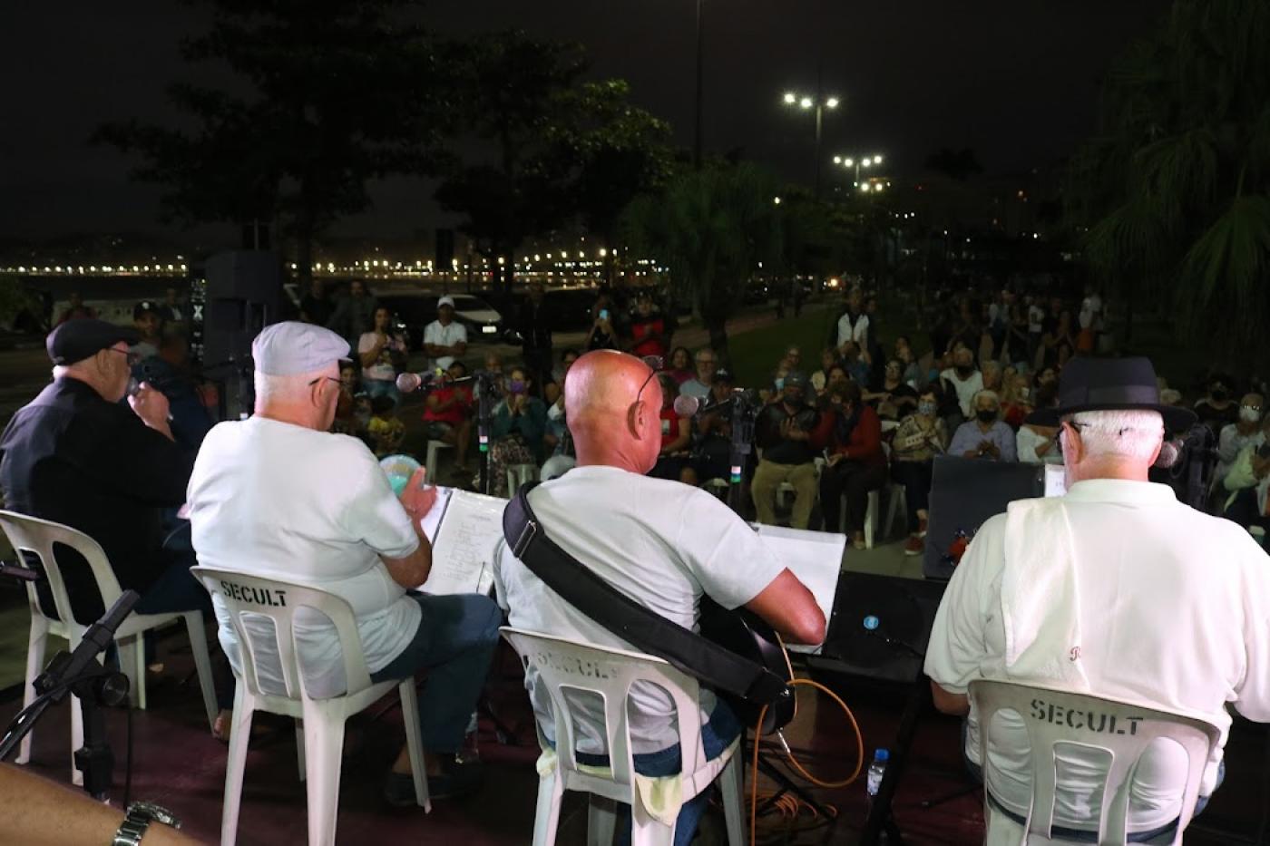 artistas tocando em palco e publico acompanhando sentado em praça #paratodosverem